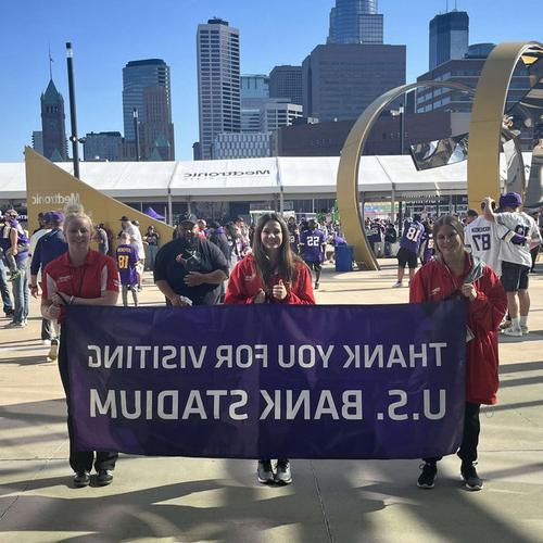 Three SCSU Swim & Dive team members smile while holding a banner at U.S. Bank Stadium